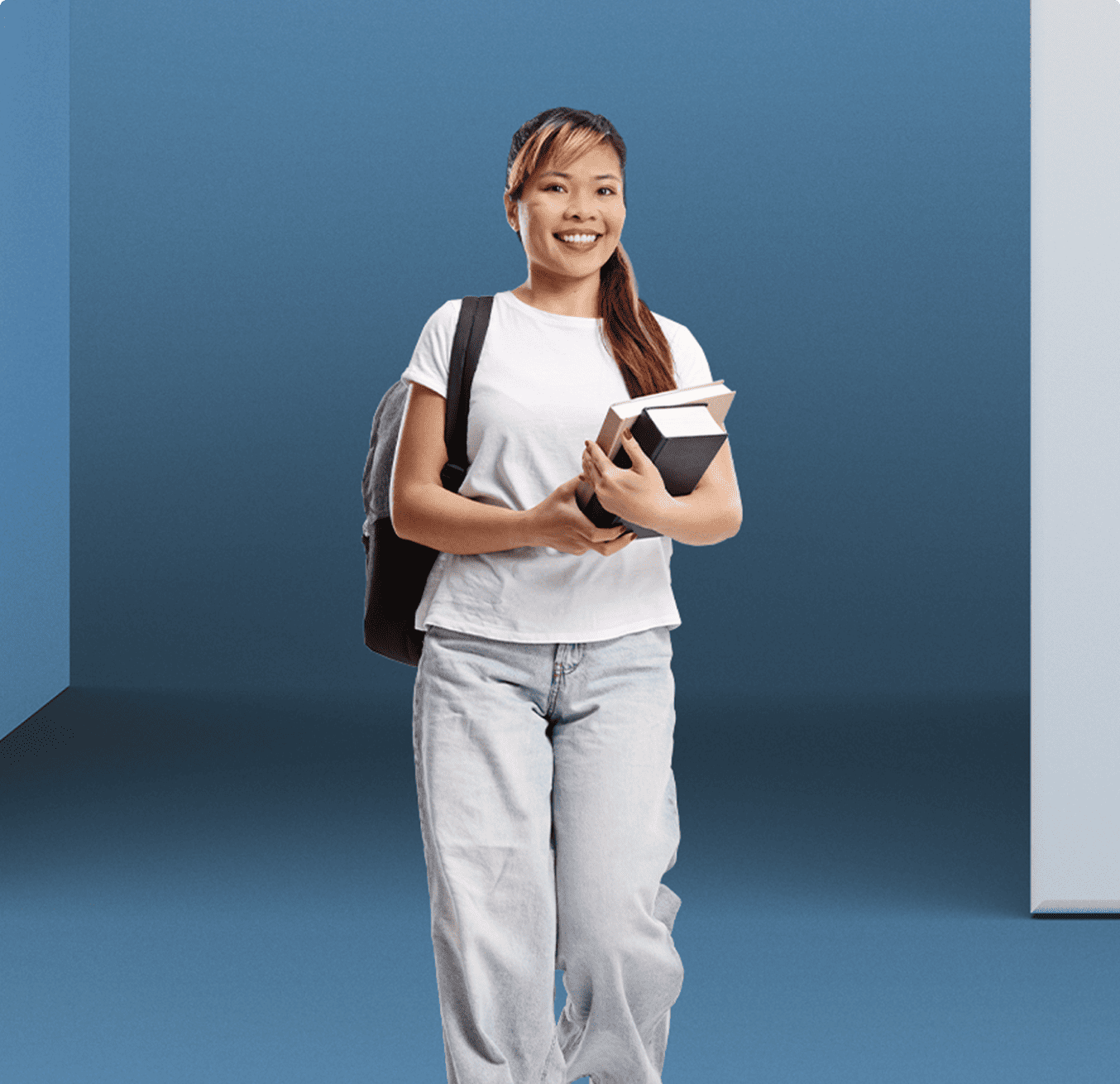 A young woman wearing a backpack and carrying textbooks smiles as she walks towards the camera.